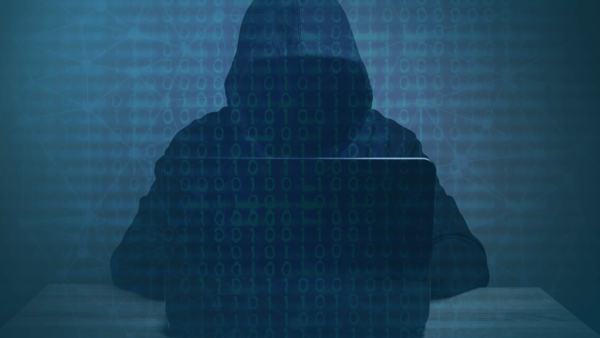 A hooded person sitting the dark on a laptop, shadows obscuring their face. Binary code overlayed across the image.
