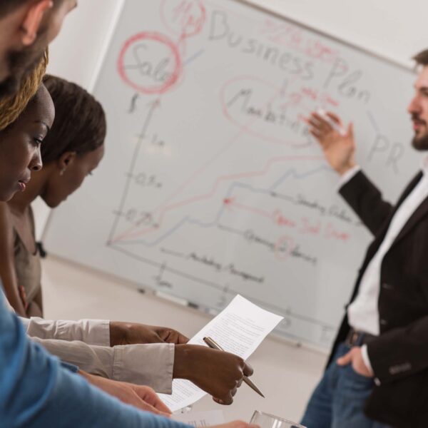 A group of people having a meeting with a man standing in front writing on a whiteboard.